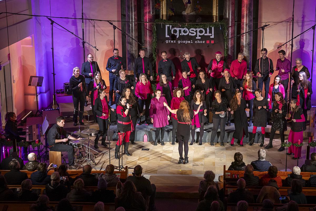 Unter leuchtend violettem Licht betritt der Grazer Gospelchor unter der Leitung eines leidenschaftlichen Dirigenten die Bühne, während das Publikum gebannt im Vordergrund sitzt.