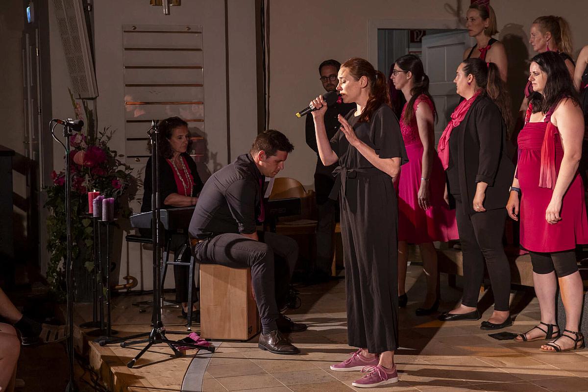 Auf der Bühne singt eine Frau in Schwarz gefühlvoll in ein Mikrofon, begleitet von einem sitzenden Mann, der ein Instrument spielt, während im Hintergrund die Mitglieder des Grazer Gospelchors in Pink wunderschön harmonieren.