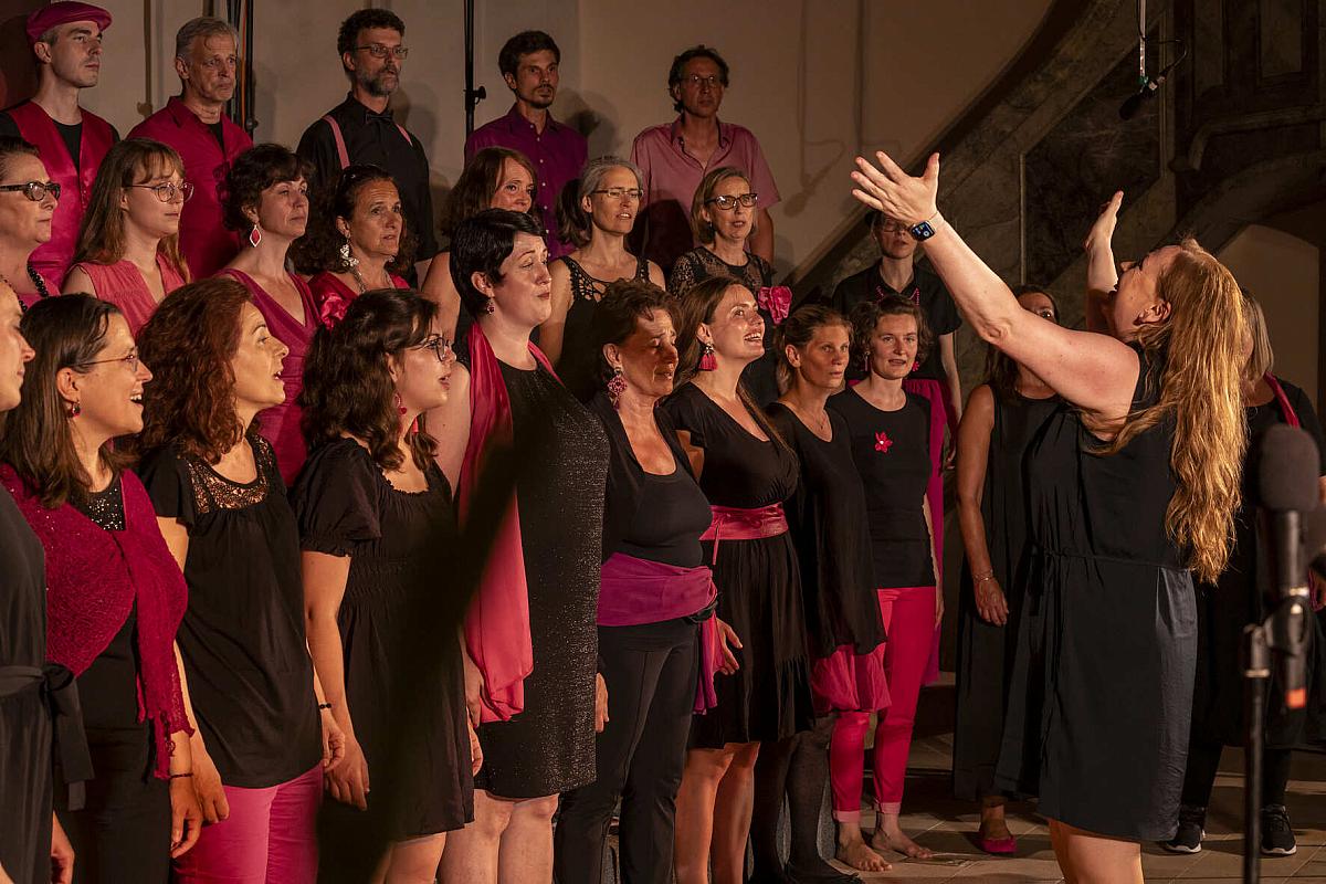 Der in Schwarz und Pink gekleidete Grazer Gospelchor tritt leidenschaftlich auf, während der Dirigent ihn mit Enthusiasmus und hoch erhobenen Armen in einer bezaubernden Innenraumkulisse leitet.