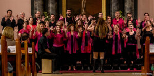 Der in Schwarz und Pink gekleidete Grazer Gospelchor tritt mit erhobenen Händen vor einem Gemälde auf. Vor ihnen steht ein Dirigent, der die harmonische Darbietung gekonnt leitet.