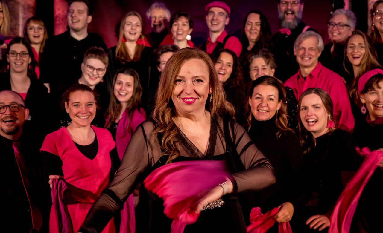 Eine Gruppe von Menschen in schwarz-rosa Outfits posiert zusammen, einige lächeln und andere halten rosa Schals. Vorne sticht eine Frau hervor, die den Graz Gospel Chor leitet und mit einem selbstbewussten Lächeln direkt in die Kamera blickt.