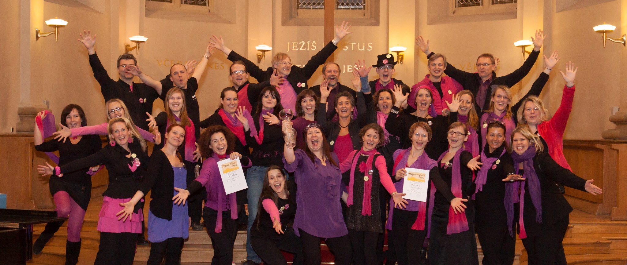 Eine fröhliche Gruppe farbenfroh gekleideter Mitglieder des Grazer Gospelchors posiert mit erhobenen Armen in einer Kirche und hält stolz ihre Zertifikate in den Händen.