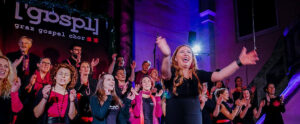 Der Grazer Gospelchor tritt energisch auf der Bühne auf, mit einer Frau als Dirigentin im Vordergrund. Schilder und buntes Licht verstärken die lebendige Atmosphäre.
