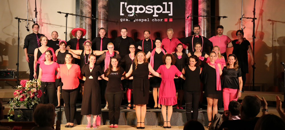 Der Grazer Gospelchor steht in schwarz-rosa Outfits vor einem Banner auf der Bühne, die Mitglieder stehen aufgereiht und halten sich an den Händen.