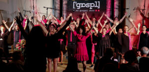 Ein in Schwarz und Pink gekleideter Chor tritt auf der Bühne auf und streckt im Gleichklang die Arme. Auf dem Banner hinter ihnen steht stolz „Graz Gospel Chor“. Im Vordergrund sind die Zuschauer zu sehen.