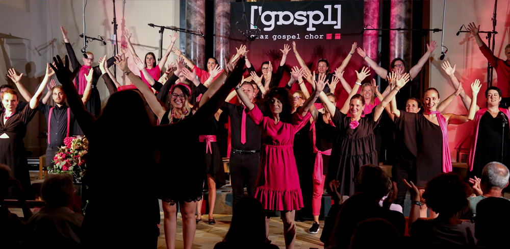 Ein in Schwarz und Pink gekleideter Chor tritt auf der Bühne auf und streckt im Gleichklang die Arme. Auf dem Banner hinter ihnen steht stolz „Graz Gospel Chor“. Im Vordergrund sind die Zuschauer zu sehen.