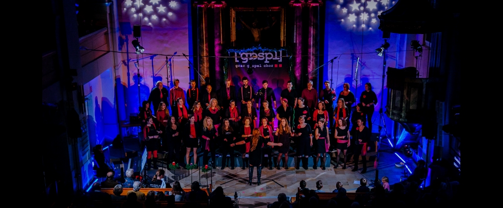 Der Graz Gospel Chor betritt die Bühne in einem schwach beleuchteten Veranstaltungsort. Männer und Frauen in Schwarz und Rot liefern eine gefühlvolle Darbietung. Musikanlage und Bühnenlicht ergänzen die bezaubernde Atmosphäre.