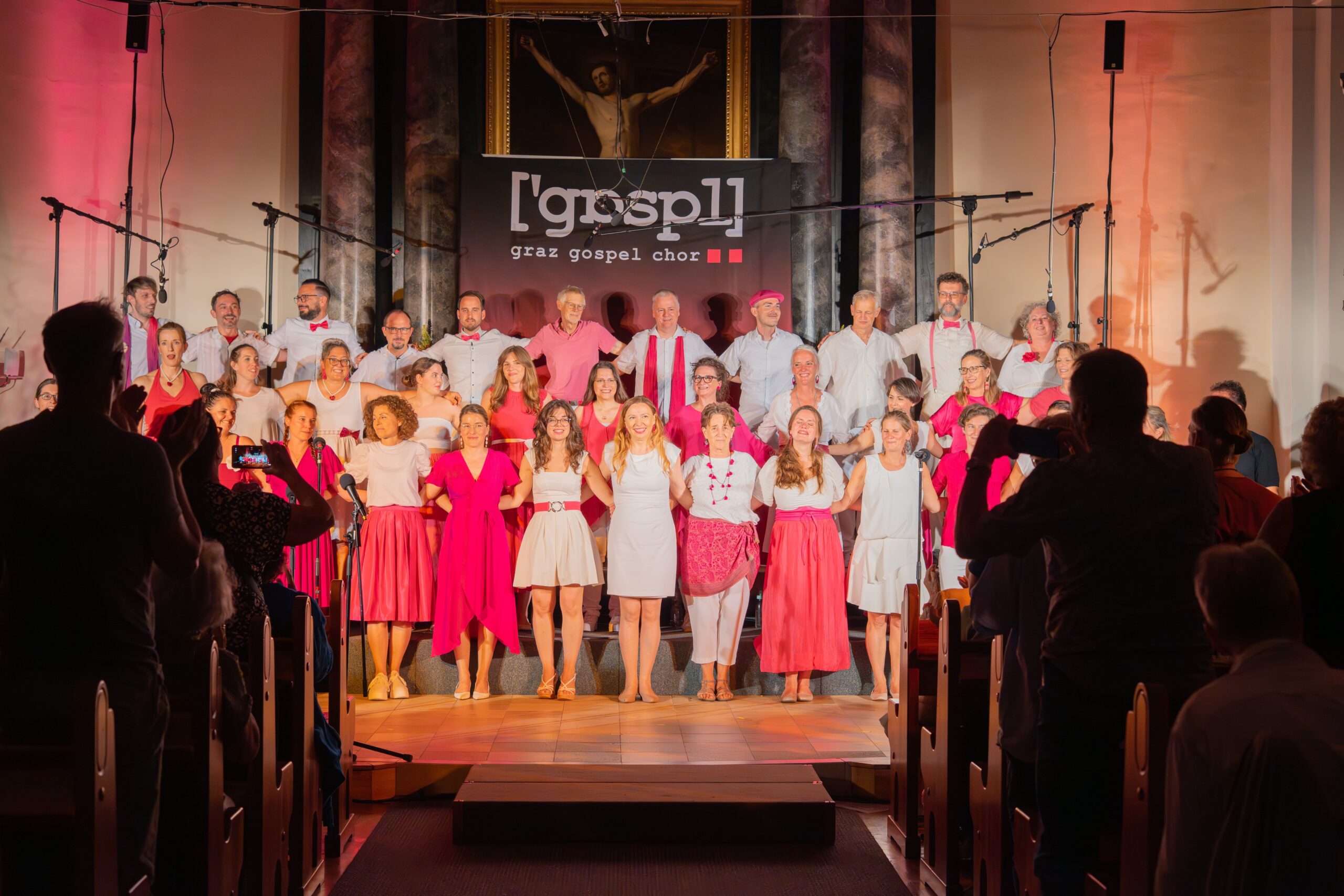 Ein großer Gospelchor steht auf der Bühne, gekleidet in koordinierten roten, rosa und weißen Outfits, und posiert für ein Gruppenfoto vor einem Publikum in einer Kirche.
